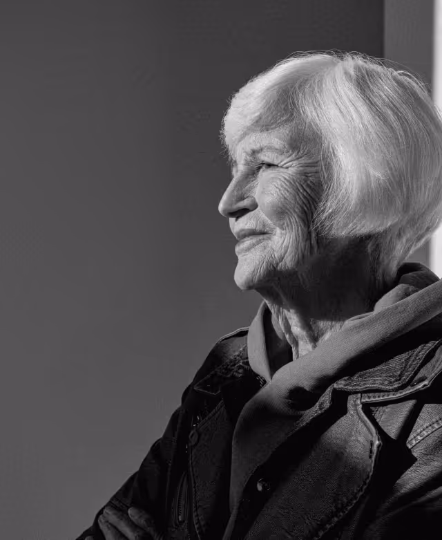 Black and white portrait of an elderly woman with white hair looking thoughtfully to the side, wearing a leather jacket and scarf.
