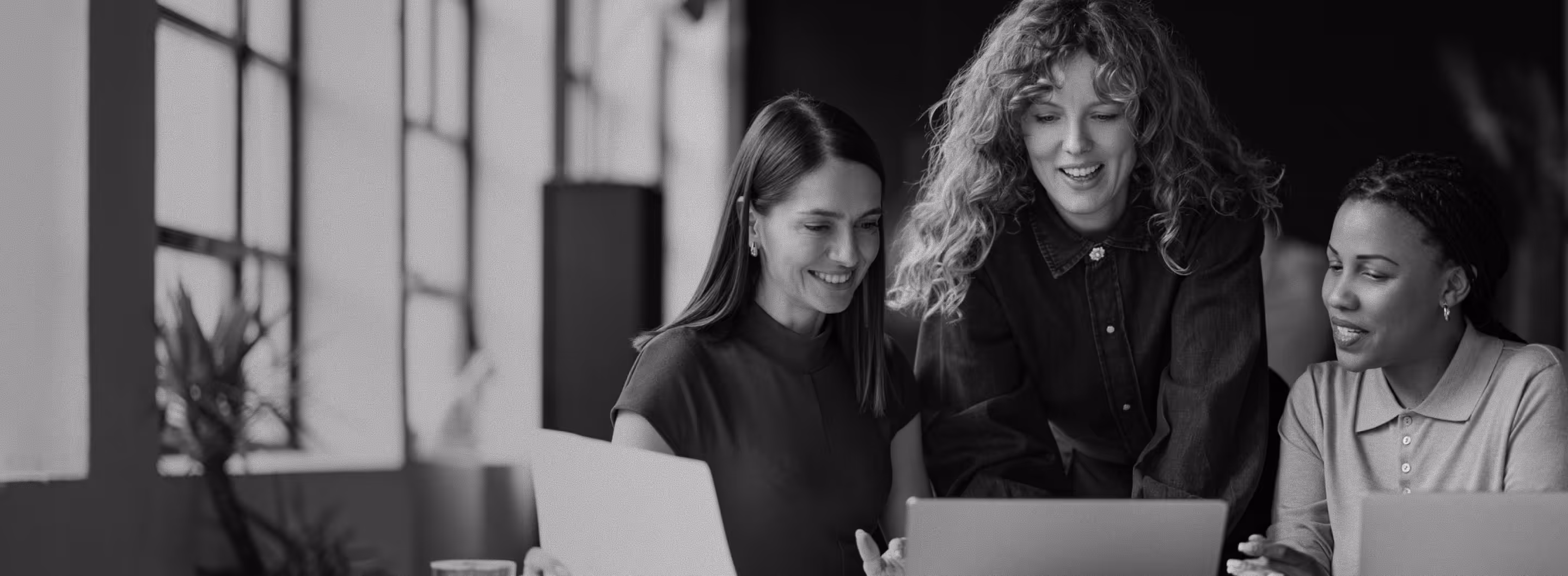 Three women gathered around laptops, smiling and engaging in discussion in a bright office space.