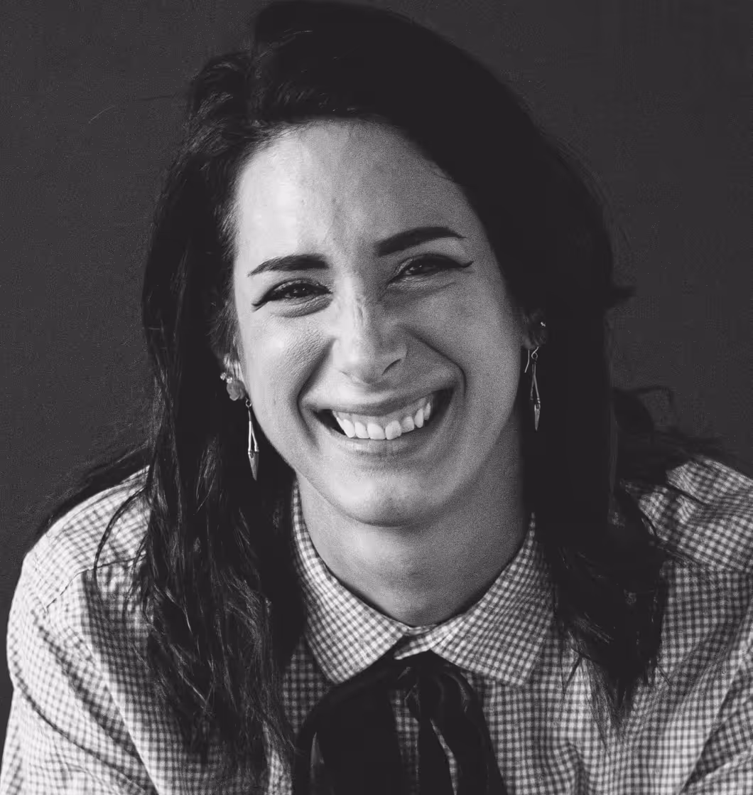 Close-up of a woman smiling with dark hair, wearing earrings and a checkered shirt with a bow tie.
