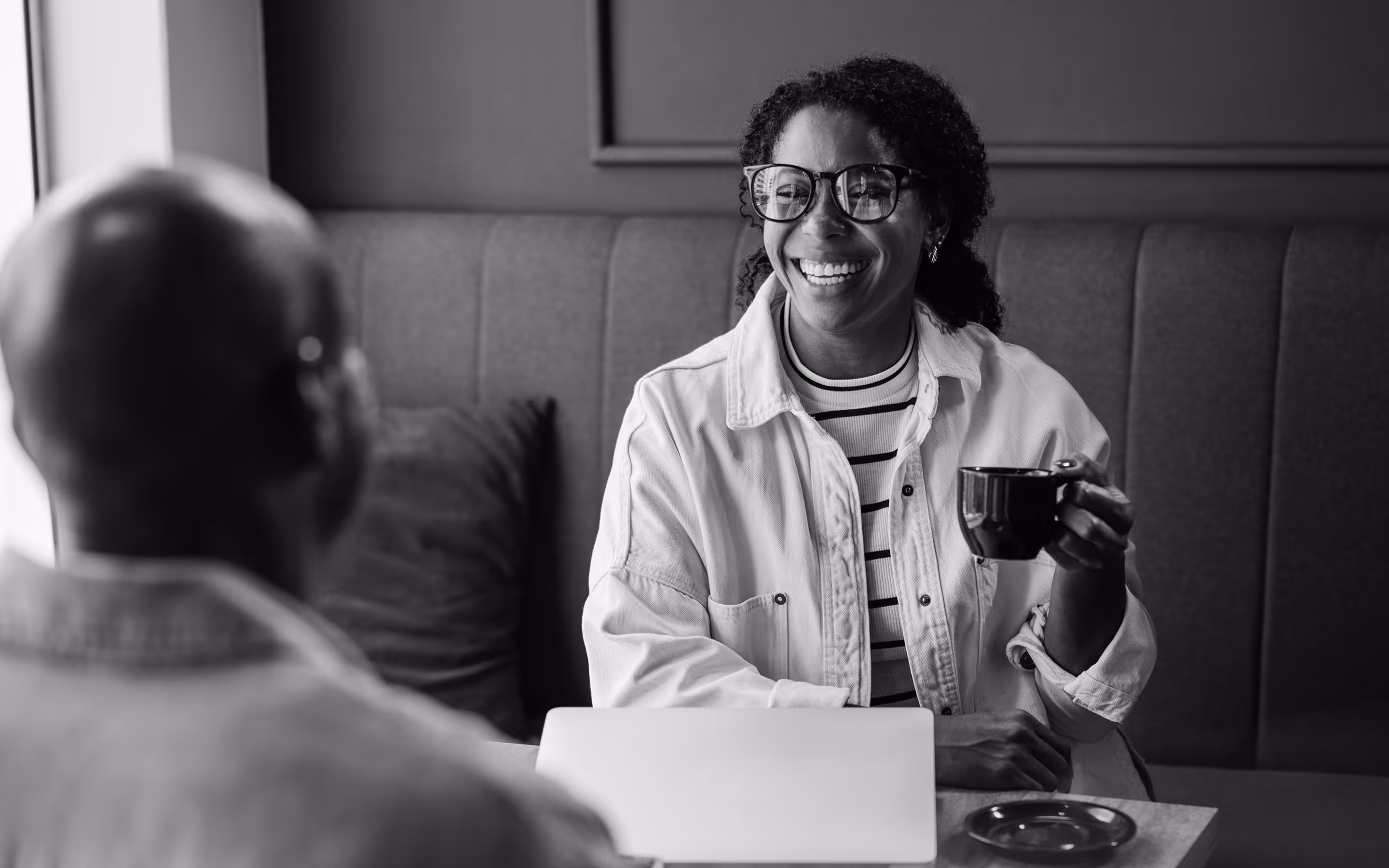 Smiling woman wearing glasses and holding a coffee cup, sitting across from a man at a table with a laptop.