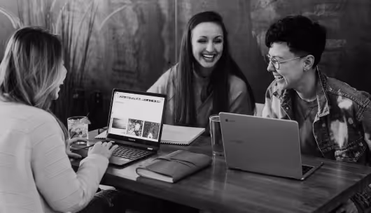 Three people sitting at a table, smiling and interacting, with laptops and notebooks in front of them.