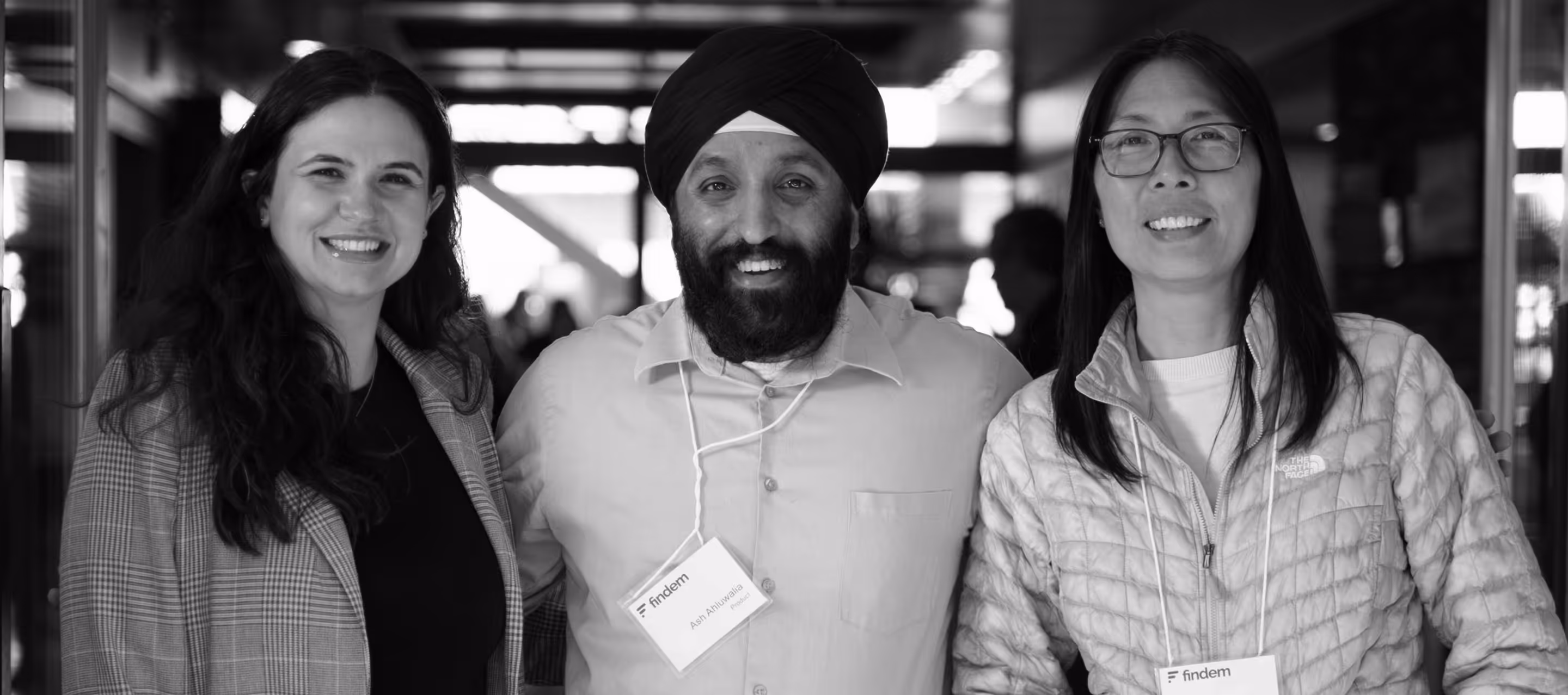 Three smiling people standing close together indoors, two women and one man wearing a turban and a badge that reads 'Ash Ahluwalia'.