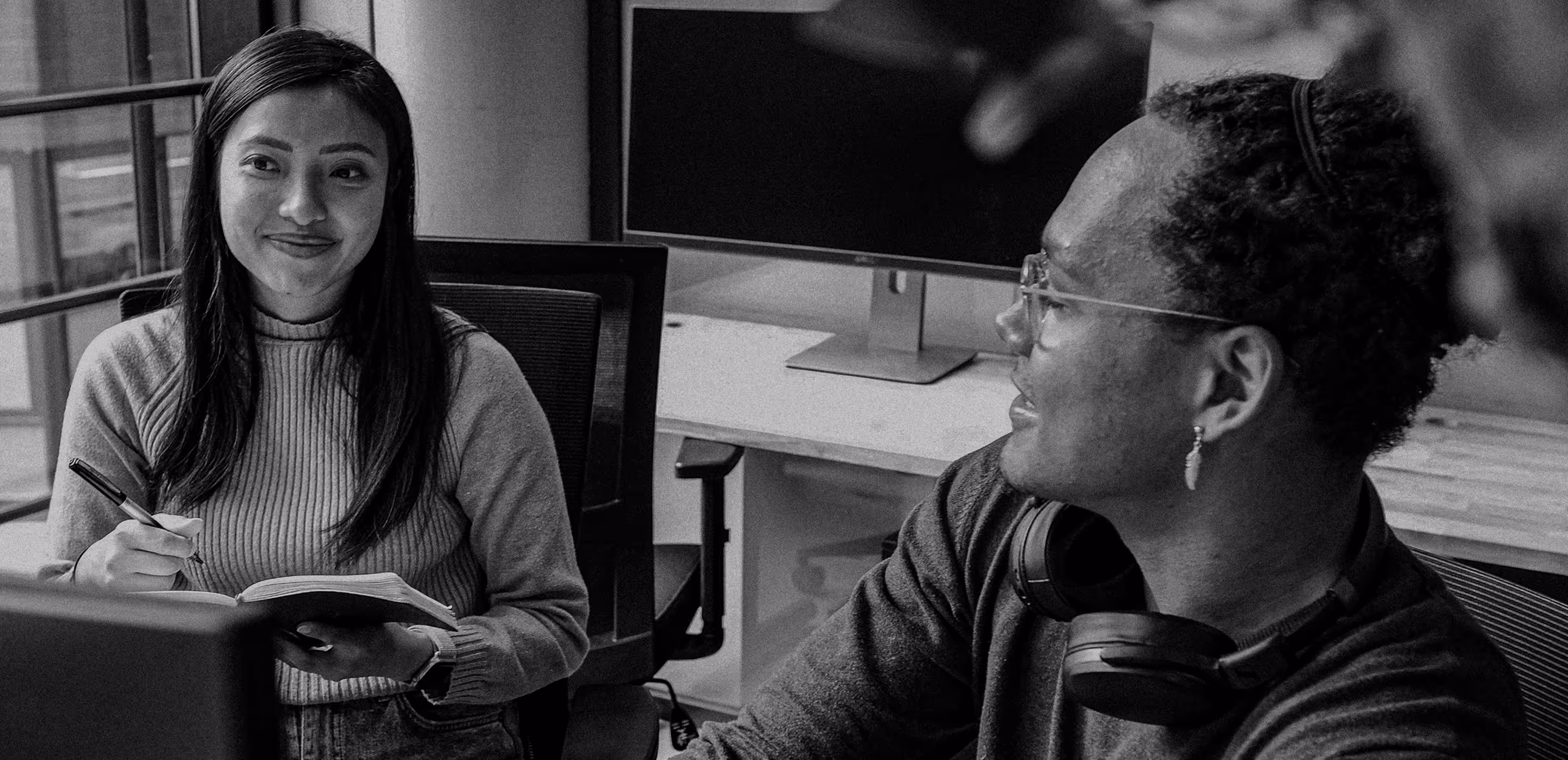 Two colleagues engaged in a conversation at a workspace, one taking notes and the other wearing headphones around the neck.