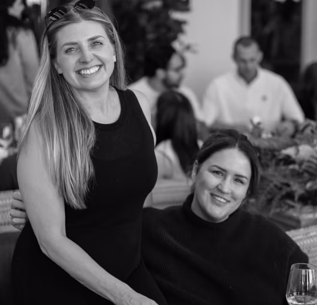 Two women smiling, one standing and one sitting, with people and a wine glass in the background.