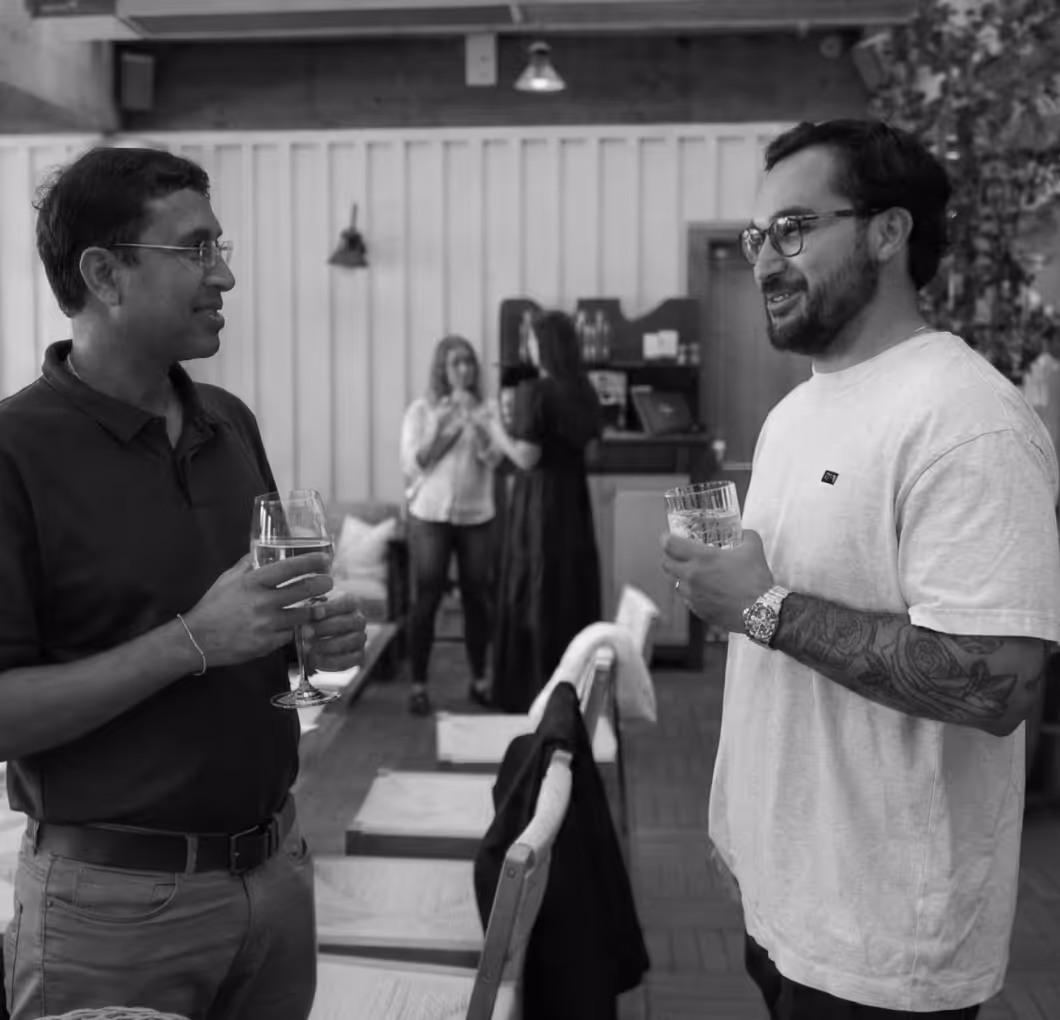Two men with glasses holding drinks and conversing indoors, with two women talking in the background.