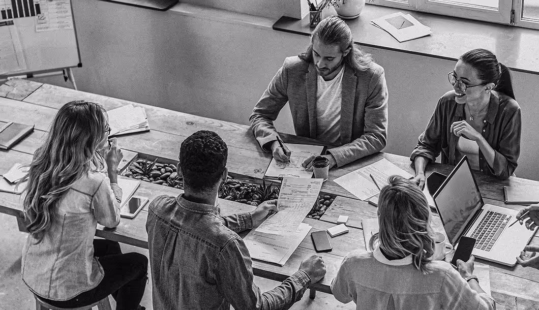 Five people collaborating around a table with documents, laptops, and phones in a meeting room.