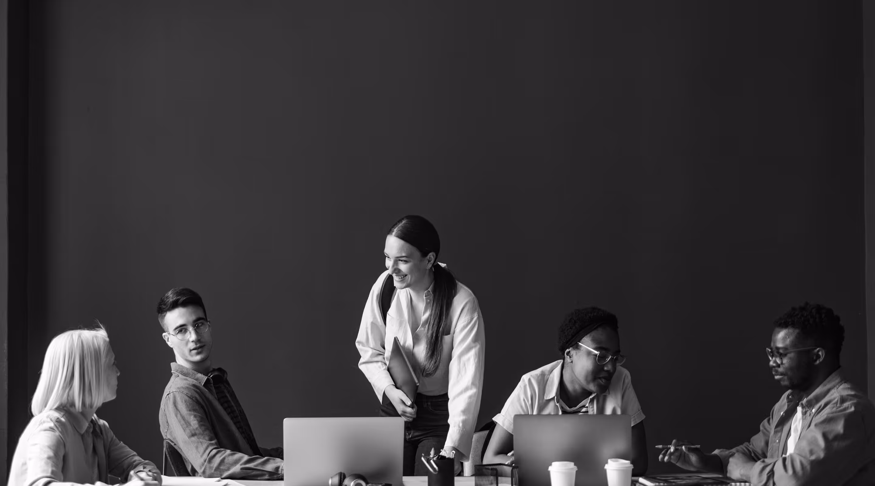 Five young professionals collaborating around a table with laptops and coffee cups in a casual meeting.