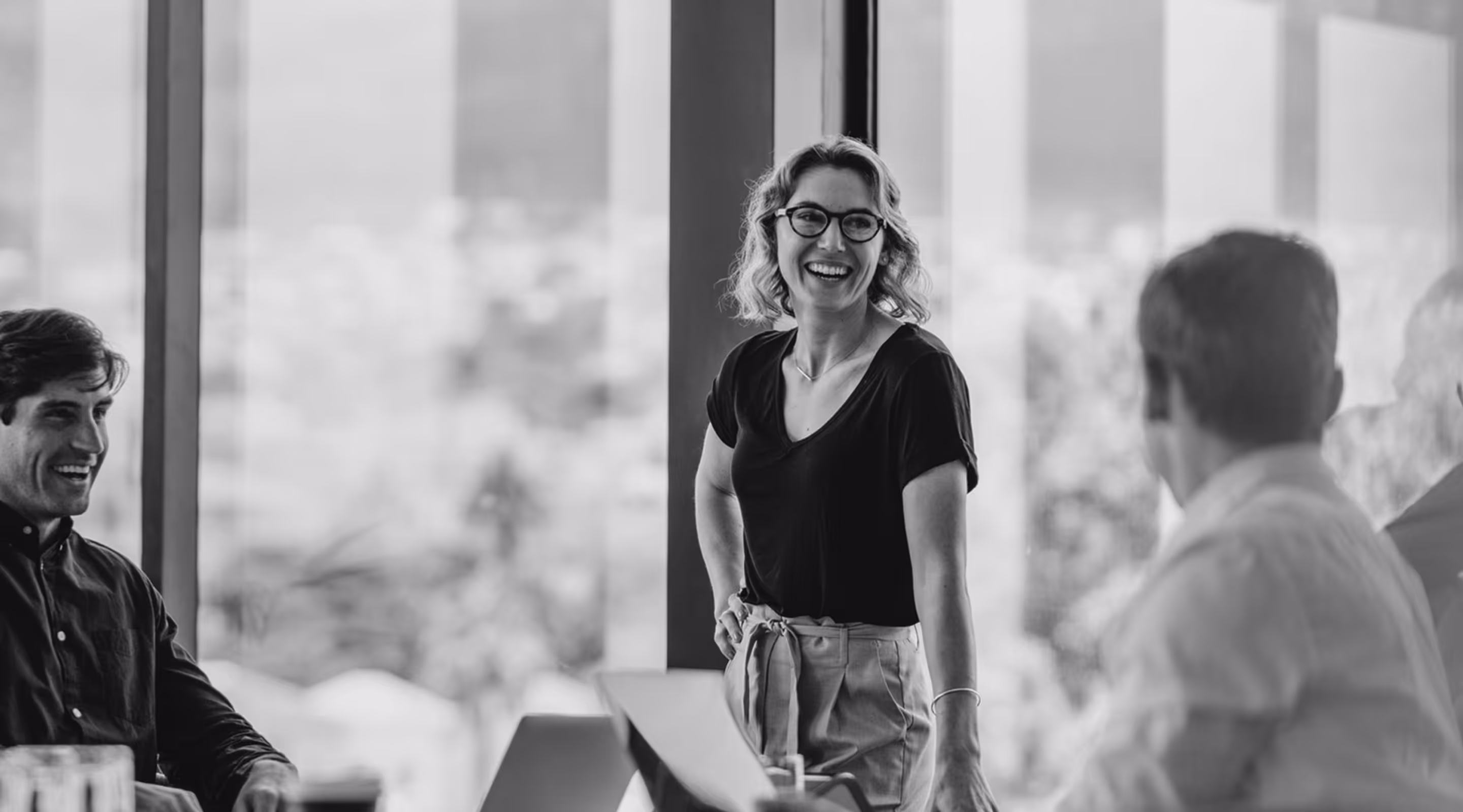 Smiling woman wearing glasses standing and talking to two seated colleagues in an office with large windows.