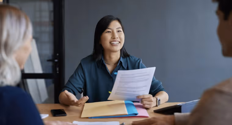 Smiling woman holding documents and gesturing during a meeting with two other people.