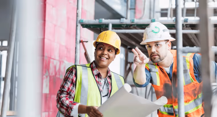 Two construction workers in hard hats and safety vests reviewing building plans on site.