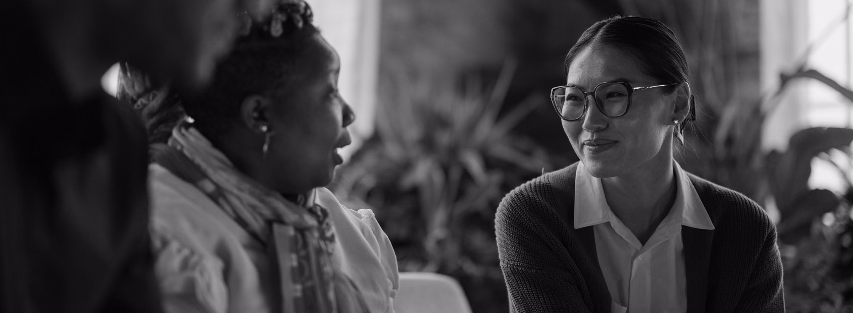 Two women engaged in a friendly conversation in a cozy indoor setting with plants in the background.